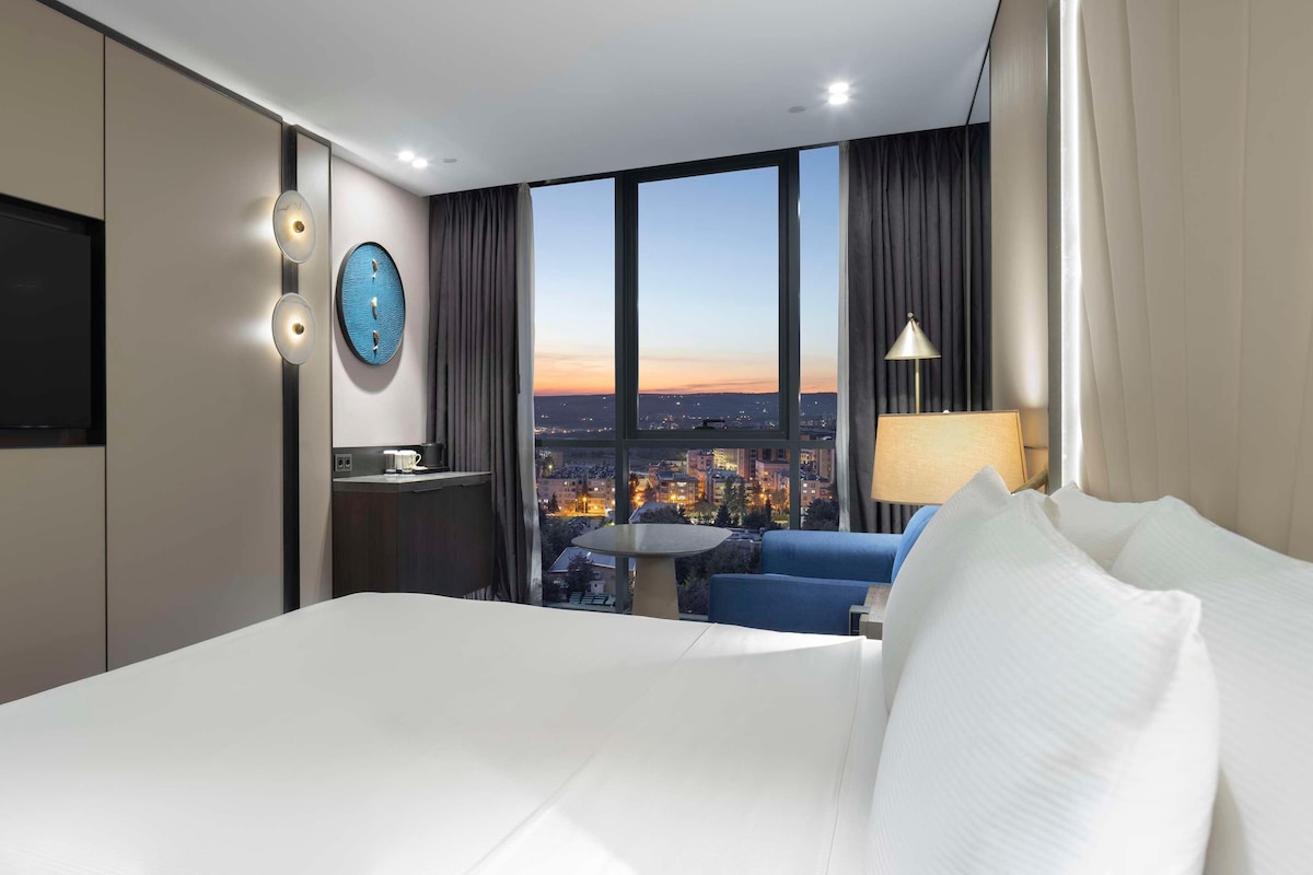 Habitación de hotel con una cama grande, televisión de pantalla plana, un reloj de pared redondo, una mesita con lámpara y vistas a la ciudad a través de amplios ventanales.