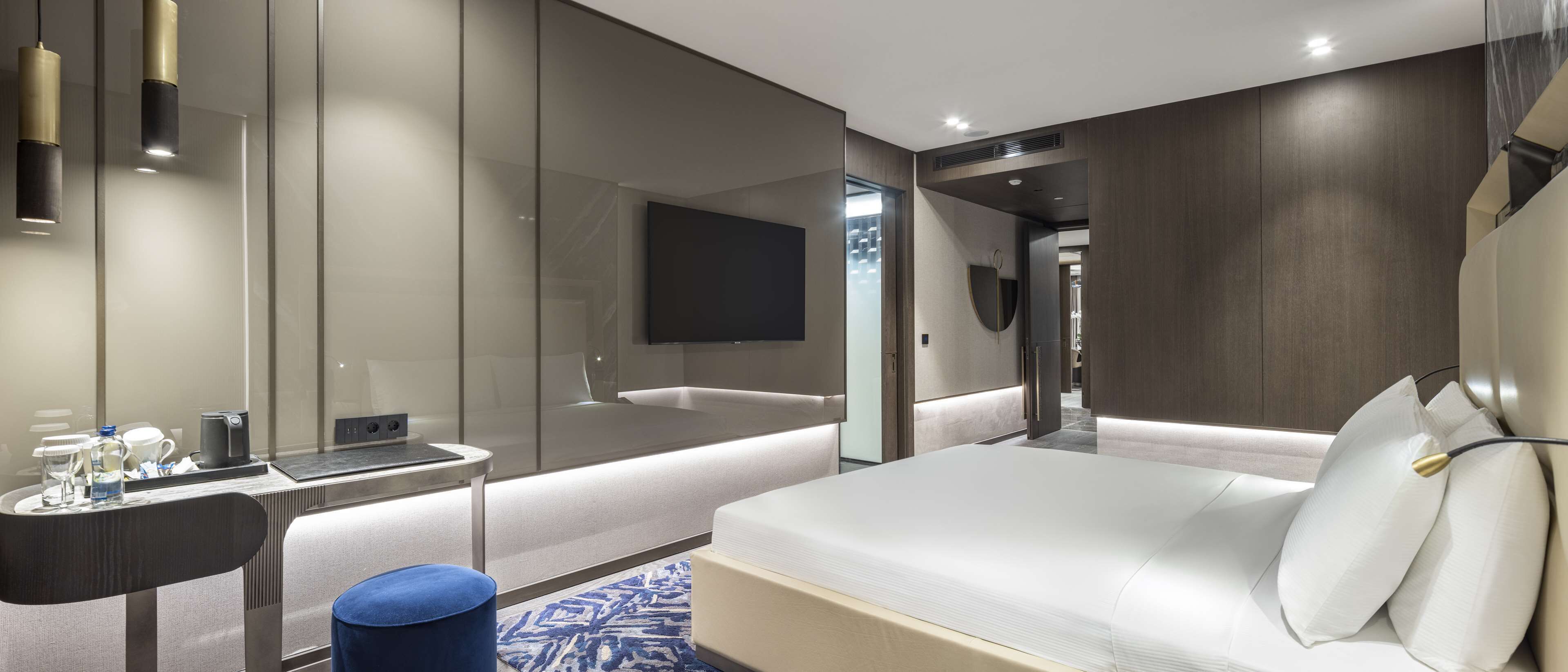 A modern hotel room with a large bed, a bedside table with a lamp, a flat-screen TV mounted on the wall, and a glass partition separating the room from the hallway.
