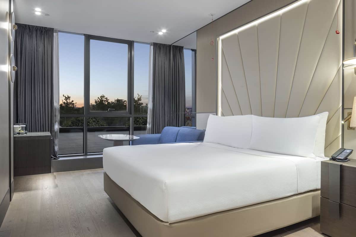 A modern hotel room with a large bed, a bedside table with a lamp, a small table, and a view of the cityscape through large windows.