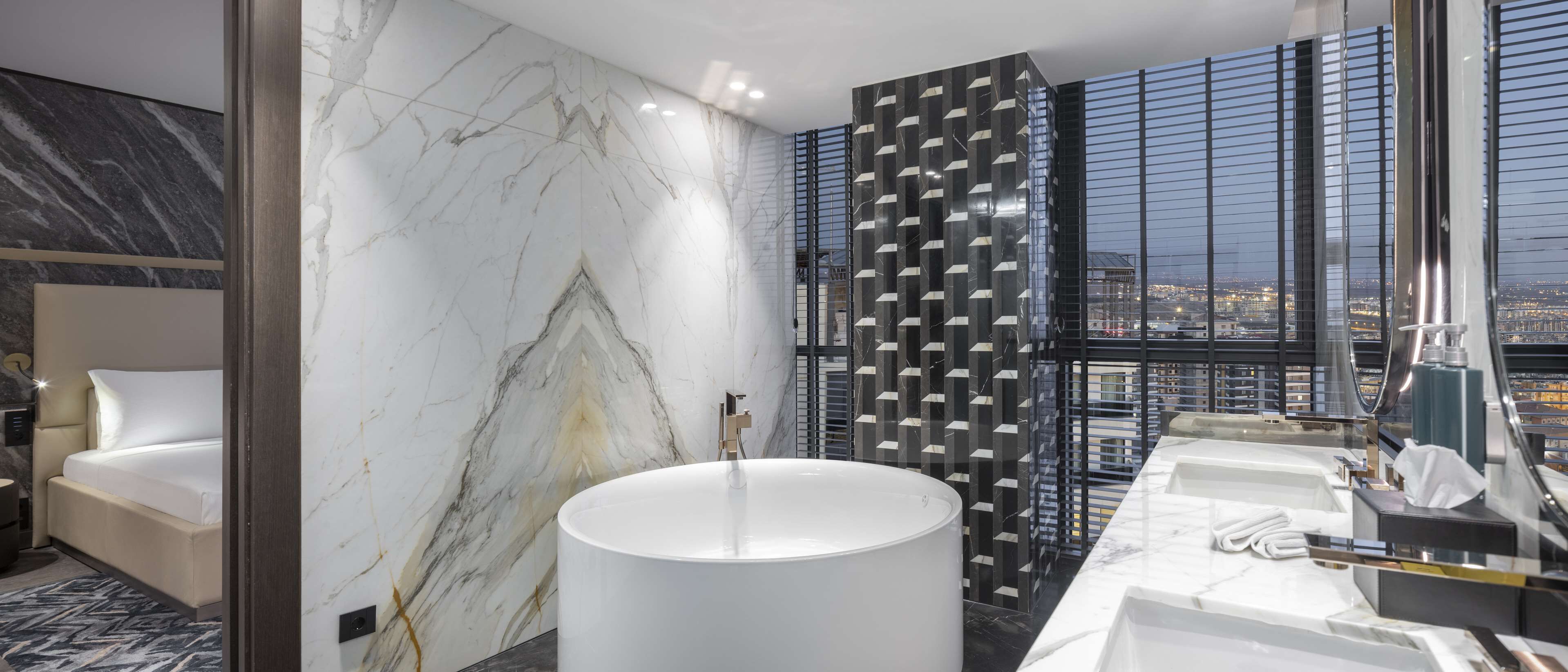 Een moderne hotelbadkamer met een grote marmeren wand, een rond vrijstaand bad en een lange wastafel met meerdere wasbakken.