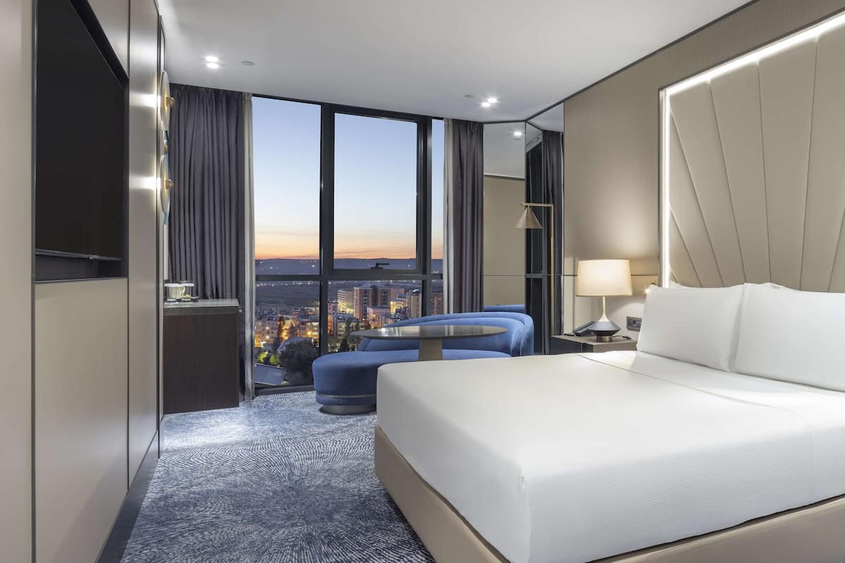 A modern hotel room with a large bed, a seating area, and a view of the cityscape through large windows.