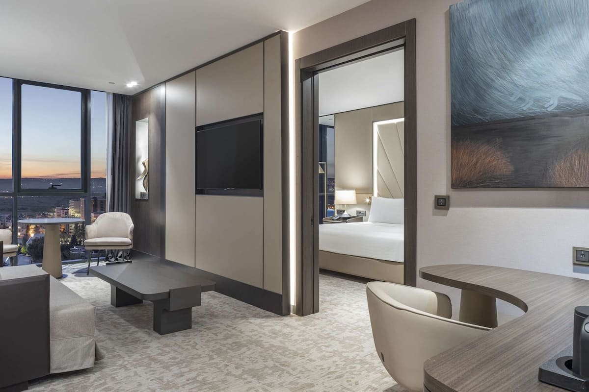 A modern hotel room with a large window offering a city view, a flat-screen TV, a sofa, a coffee table, and a desk with a lamp.