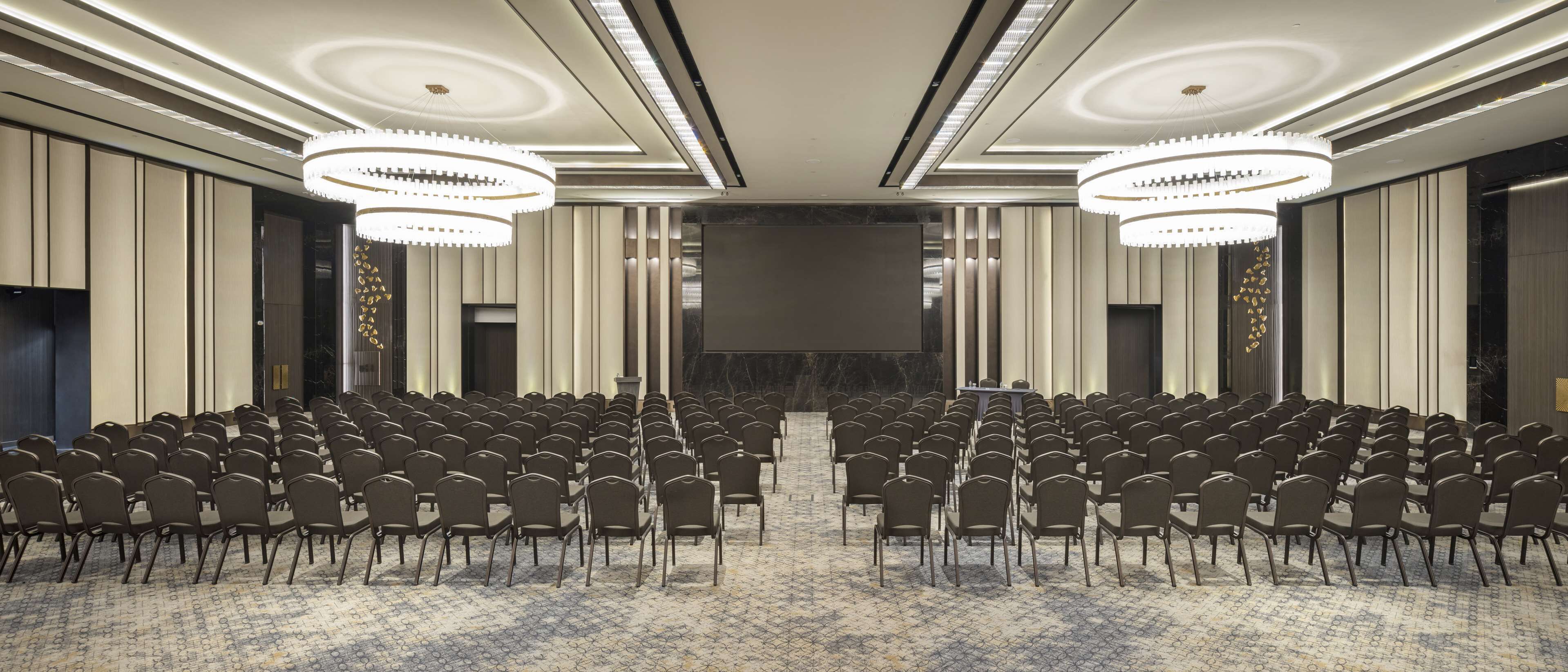 A conference room with rows of chairs, a large screen, and modern chandeliers.