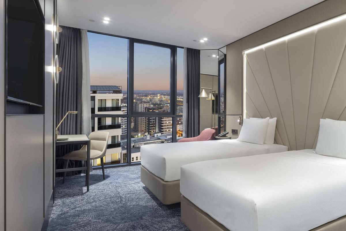 A hotel room with two beds, a desk, and a chair. The room has a large window with a city view.