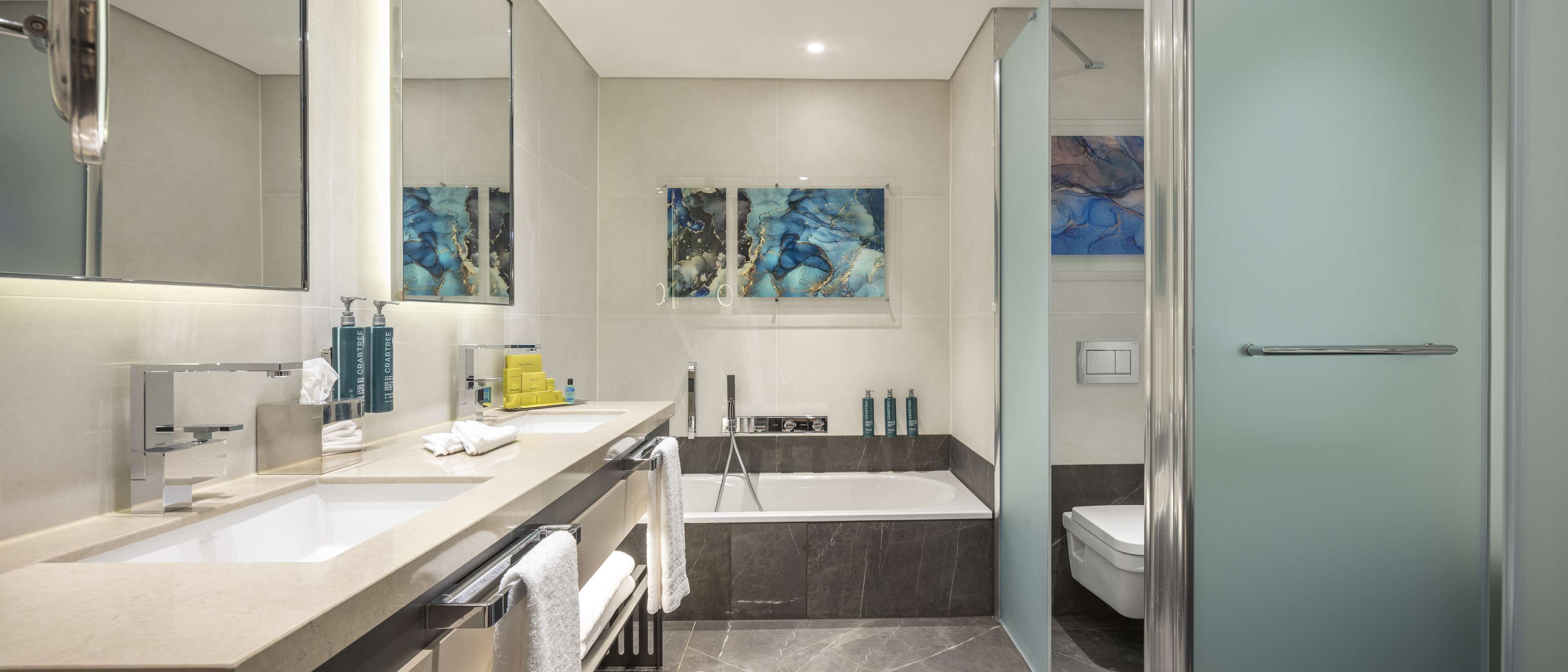 A modern bathroom with two sinks, a bathtub, and a toilet. There are towels and toiletries on the counter.