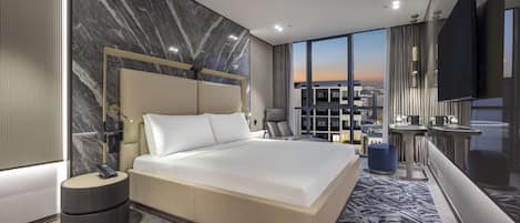A modern hotel room with a large bed, a city view, a desk with a sink, and a TV.