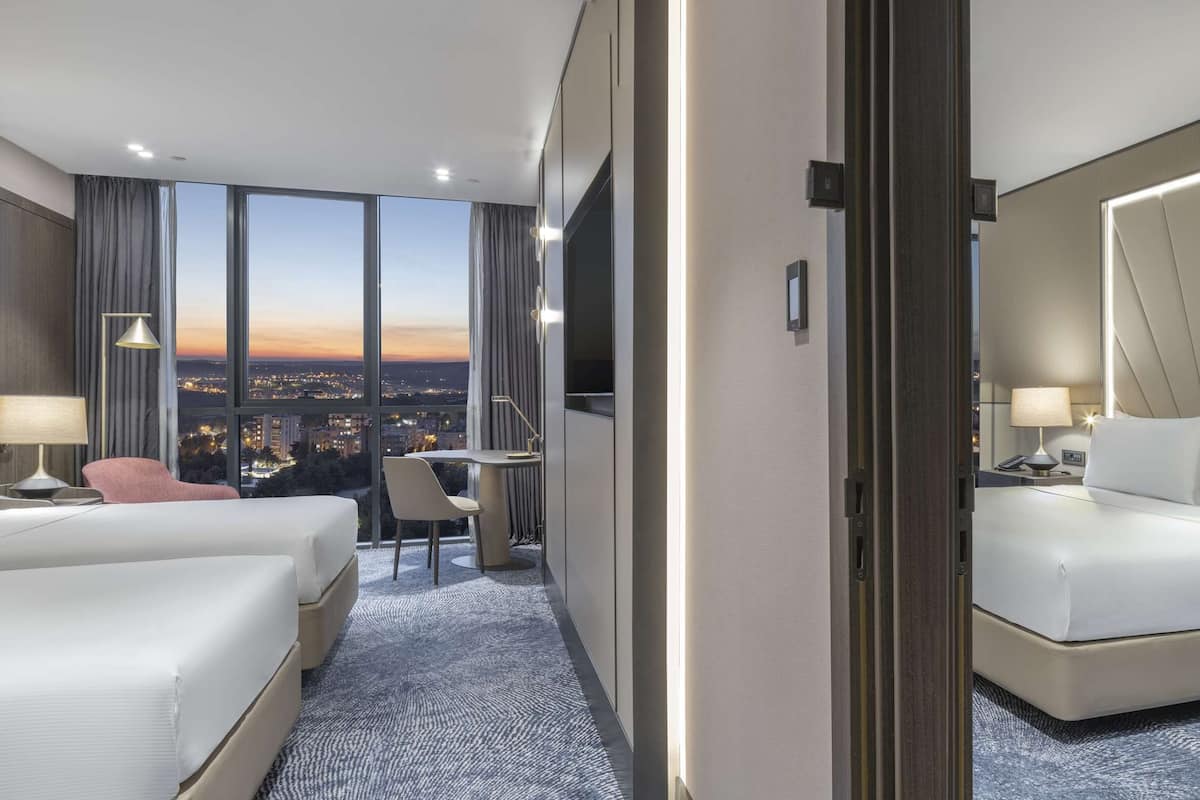 A hotel room with two beds, a desk, a chair, and a large window offering a city view.