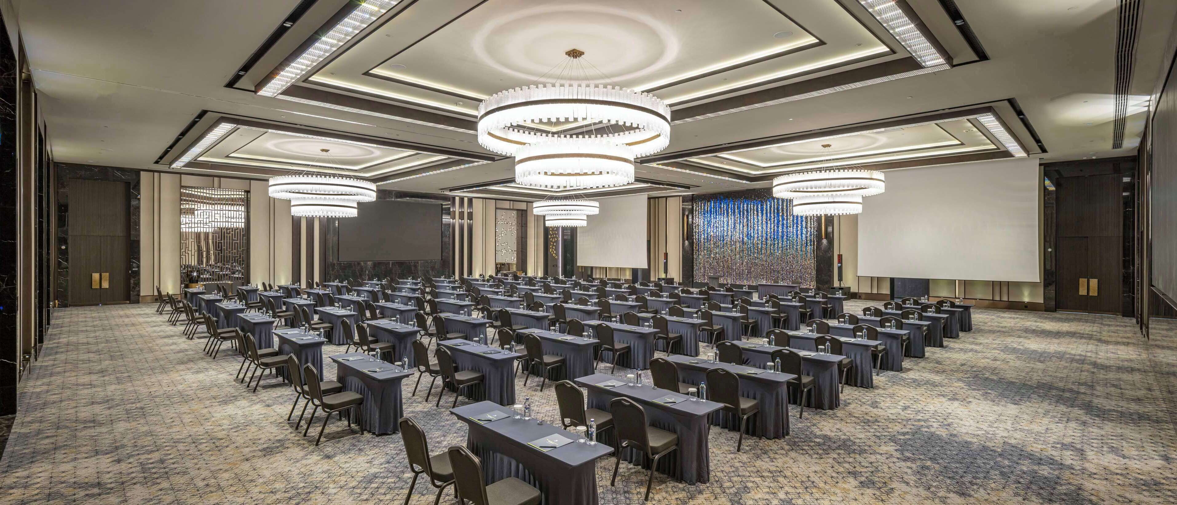 A conference room with rows of tables and chairs, large chandeliers, and a carpeted floor.