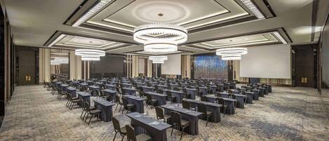 A conference room with rows of tables and chairs, large chandeliers, and a carpeted floor.