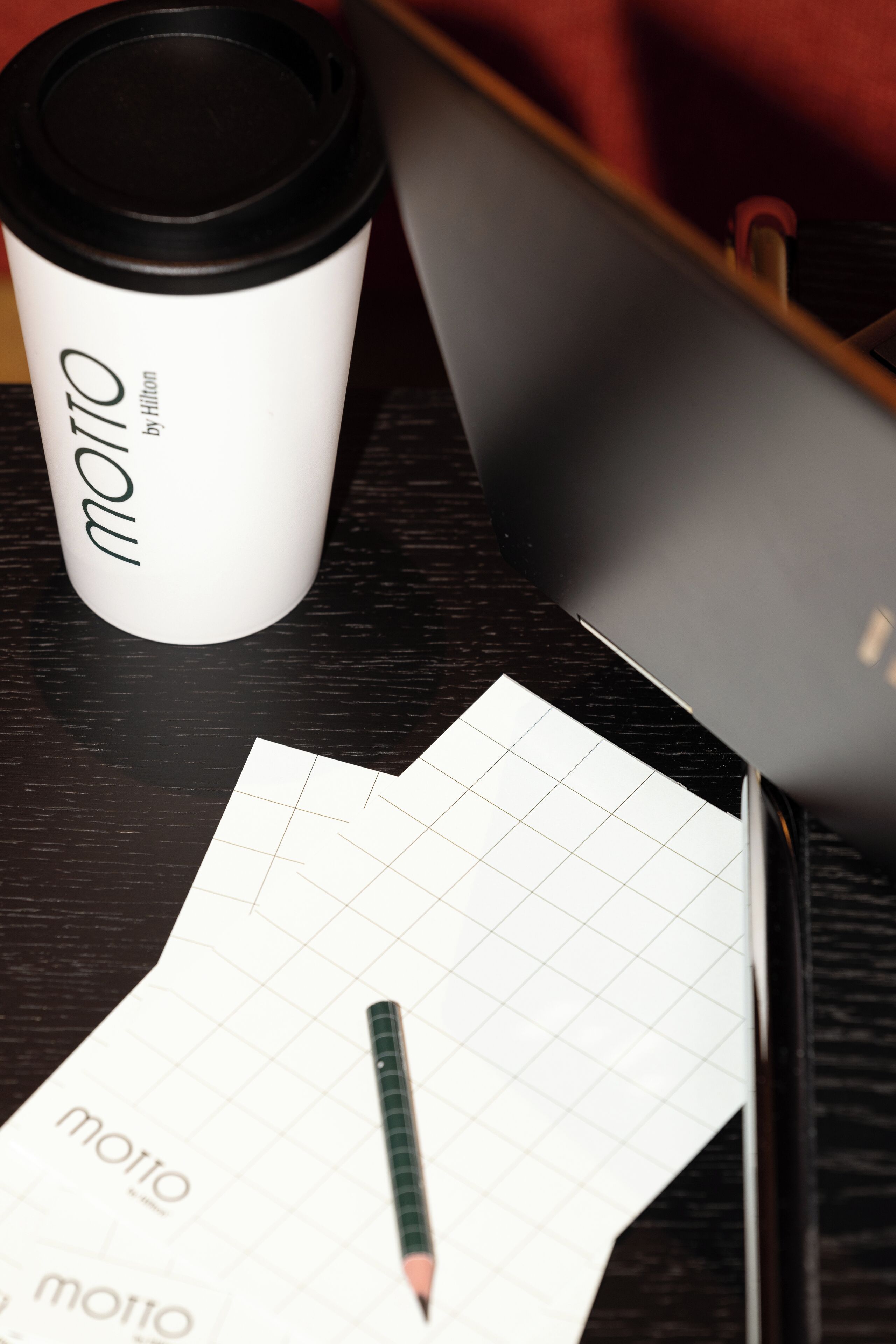 Papel blanco con líneas de cuadrícula, un lápiz y una taza de café con la etiqueta 'moto by Hilton' sobre una superficie oscura.