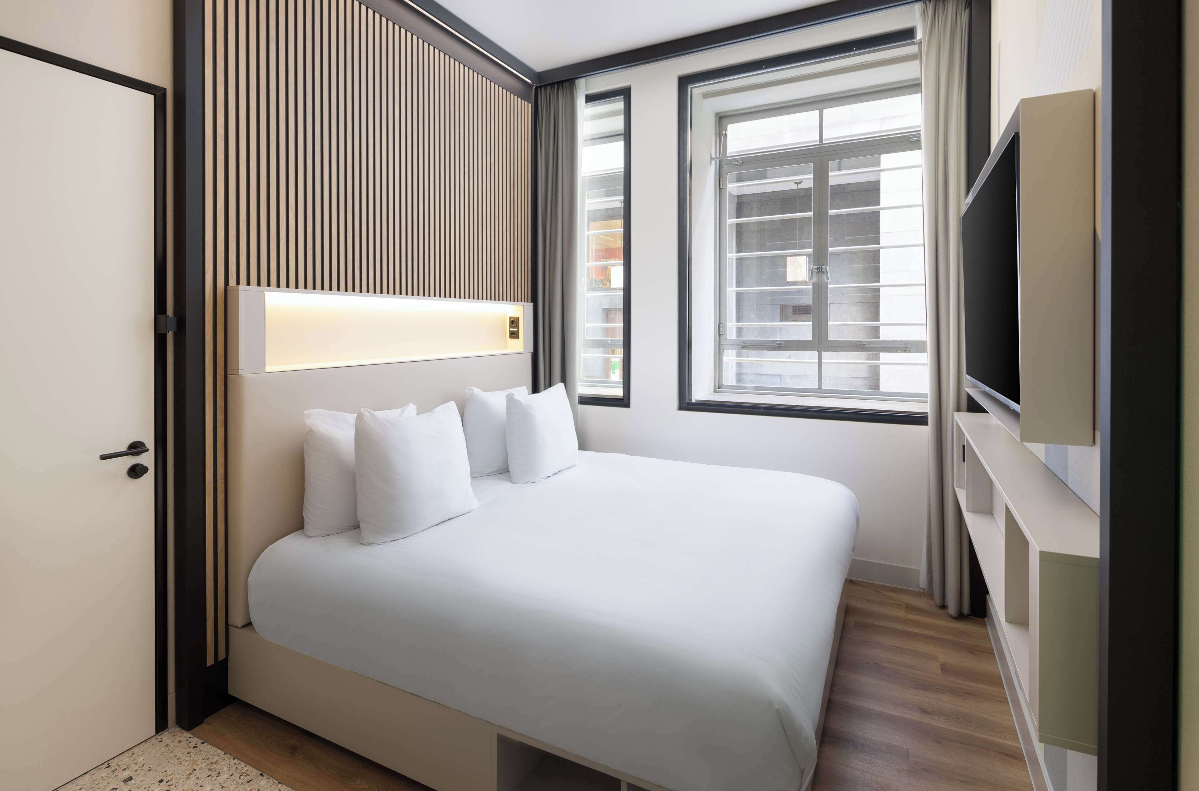 Ein modernes Hotelzimmer mit einem großen Bett, einem Fernseher und einem Fenster mit Jalousien.