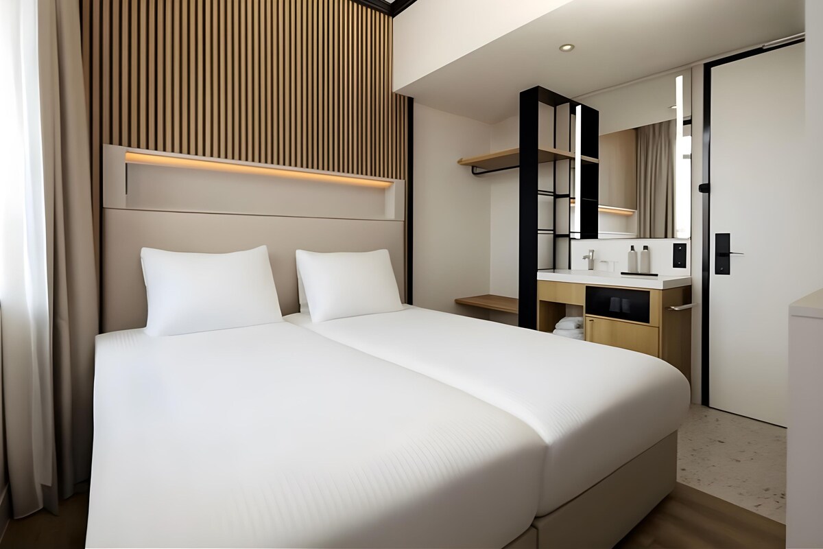 A modern hotel room with a large bed, a bedside table, a mirror, and a built-in wardrobe.