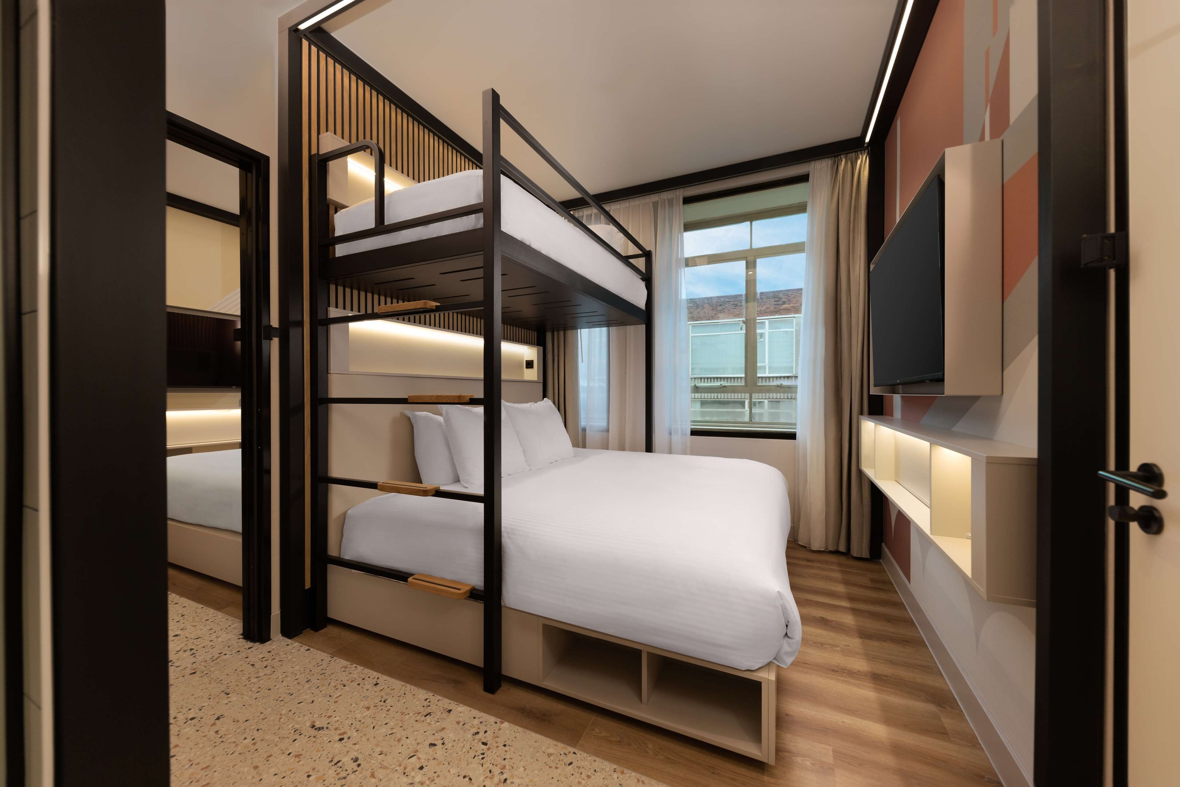 Una habitación de hotel moderna con una cama individual, una cama grande, un televisor y un ventanal con cortinas.
