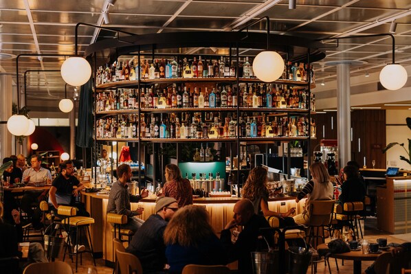 A bar area with multiple shelves of bottles, a counter, and several patrons seated at tables. - Motto by Hilton Rotterdam City Centre (Rotterdam)