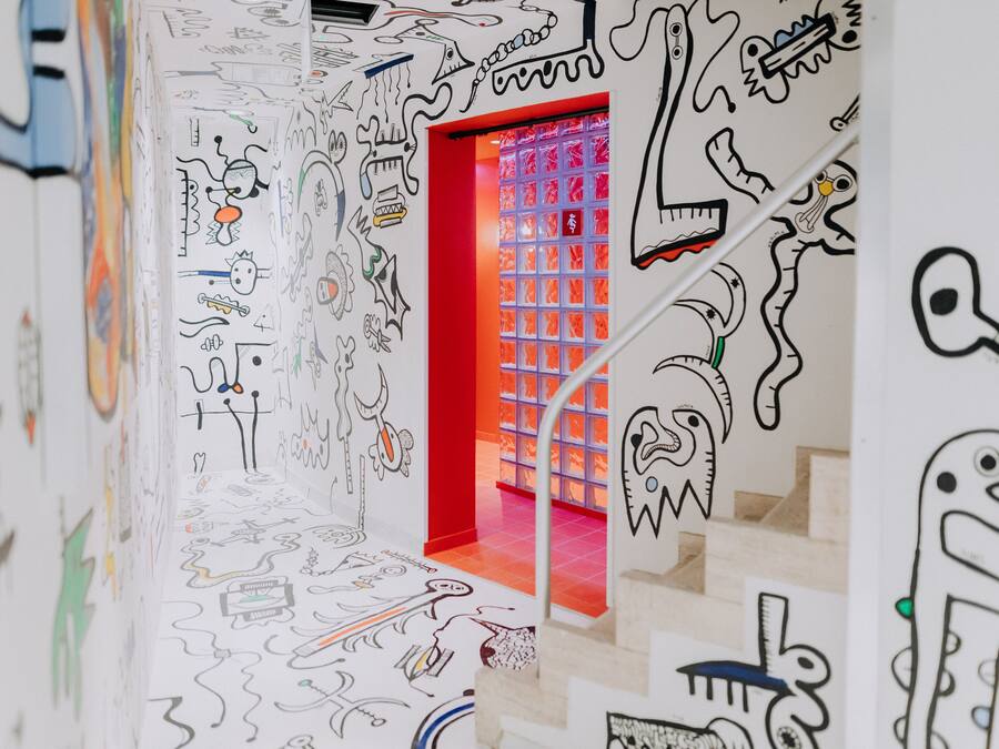 Un pasillo con paredes cubiertas de diversos garabatos y dibujos, que conduce a una habitación roja y luminosa con una ventana de diseño a cuadrícula.