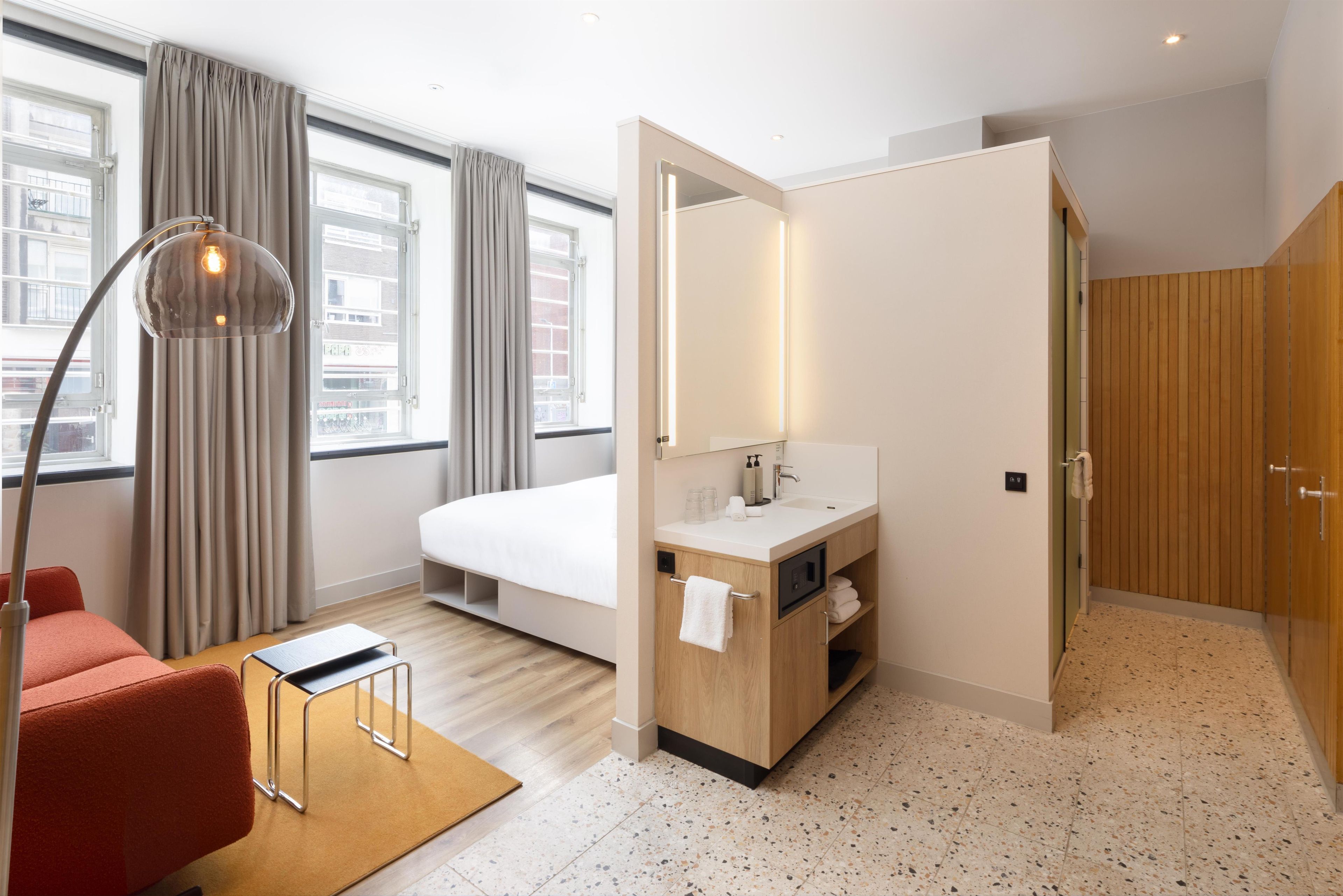 A hotel room with a bed, a bathroom with a sink and mirror, a red chair, and a floor lamp.