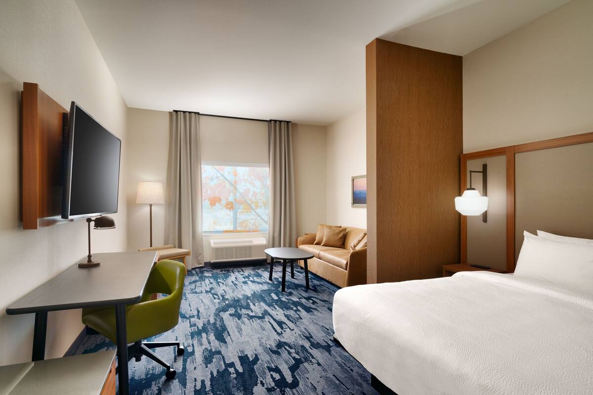 A hotel room with a bed, a desk, a chair, a sofa, a TV, and a window with curtains.