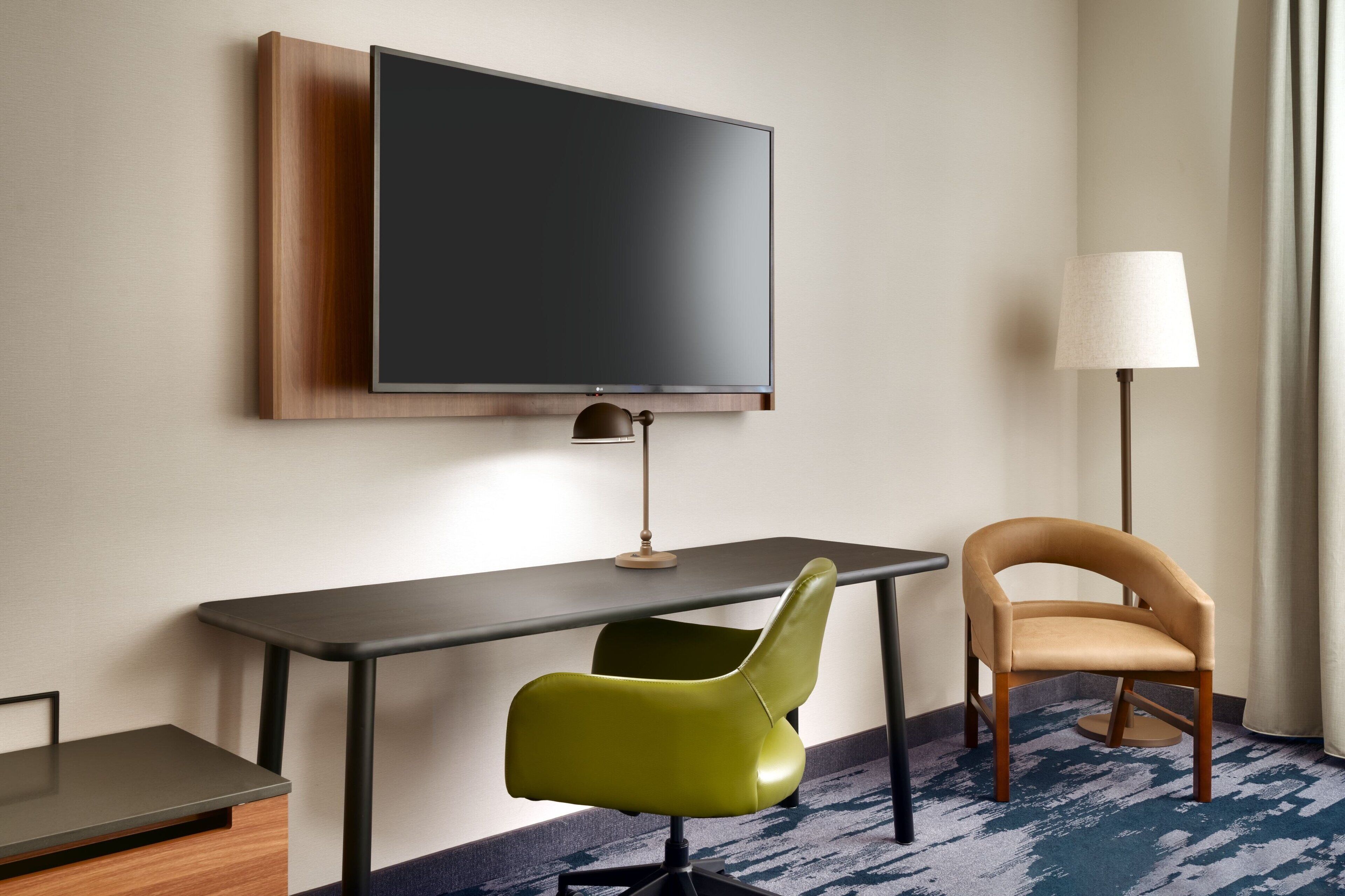 A hotel room with a flat-screen TV mounted on the wall, a desk with a lamp, a green chair, a beige armchair, and a patterned carpet.