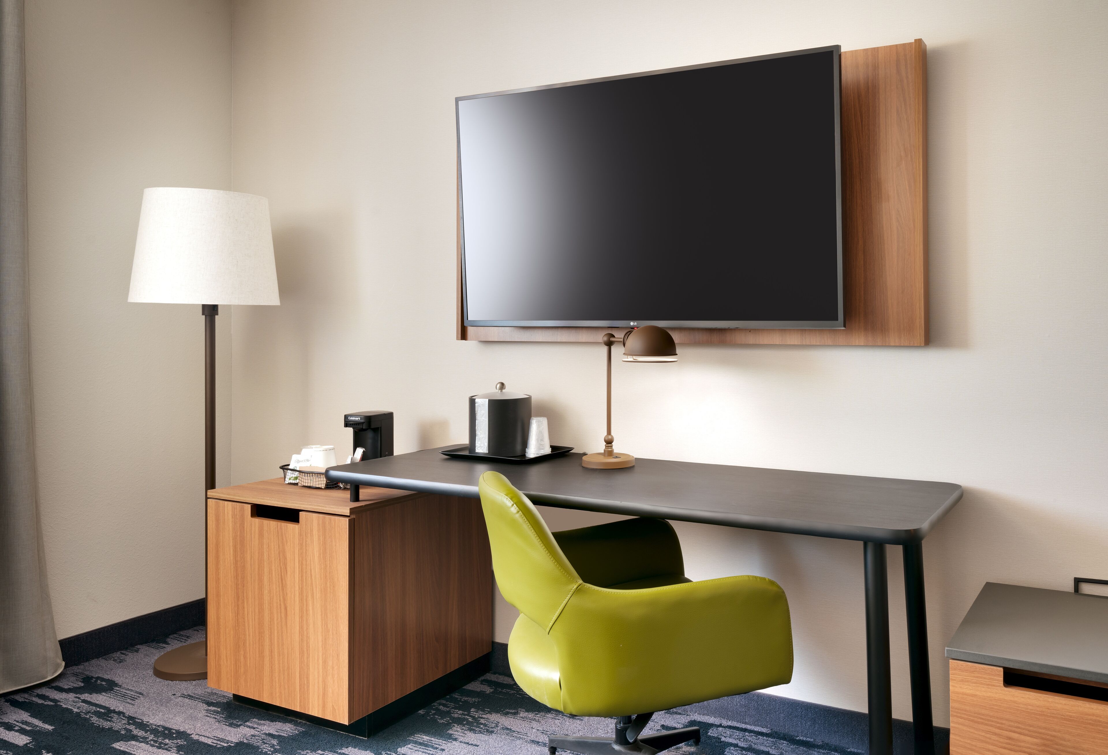 A hotel room with a flat-screen TV mounted on the wall, a desk with a lamp, a chair, and a small cabinet.