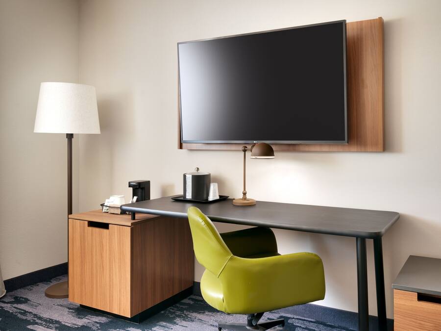A hotel room with a flat-screen TV mounted on the wall, a desk with a lamp, a chair, and a small cabinet.