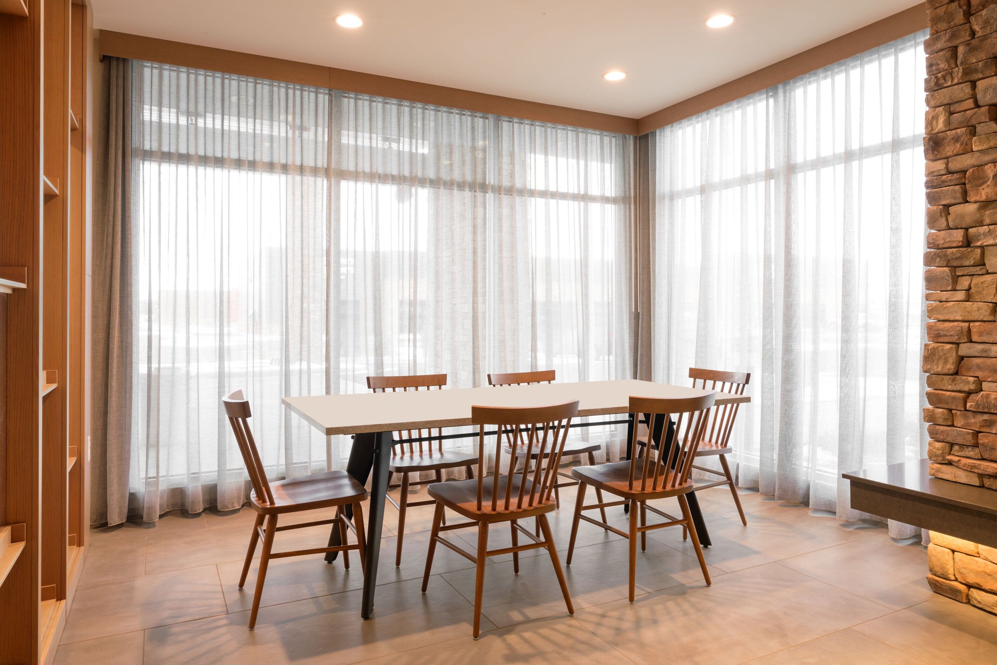 Een eetgedeelte met een grote tafel en stoelen, grote ramen met transparante gordijnen, en een wand met stenen details.