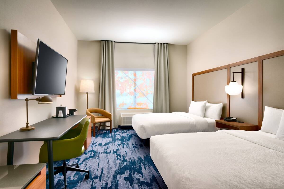 A hotel room with a desk, two beds, a TV, a lamp, and a window with curtains.