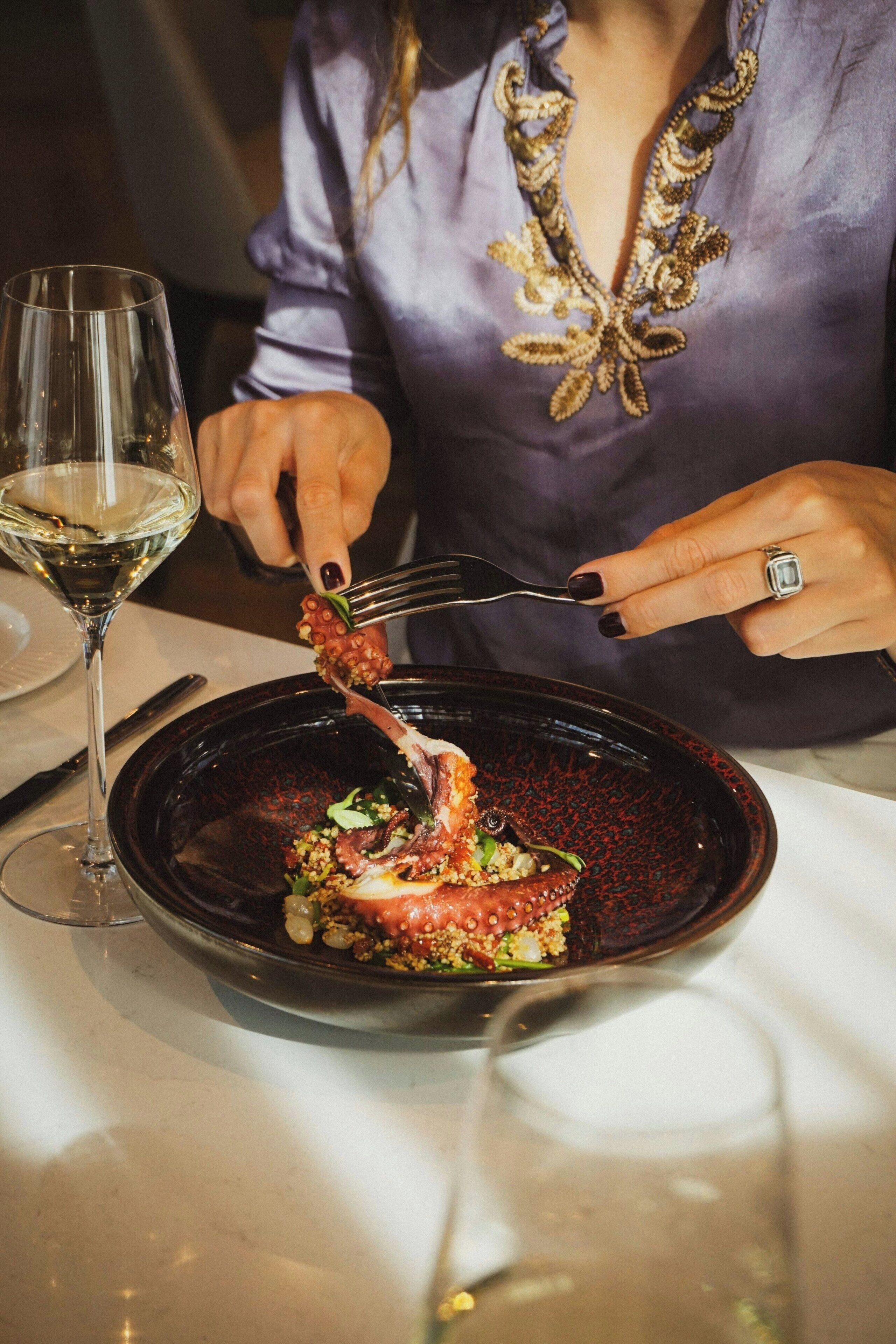 A person dining at a table with a glass of white wine, a dark plate with food, and a fork.
