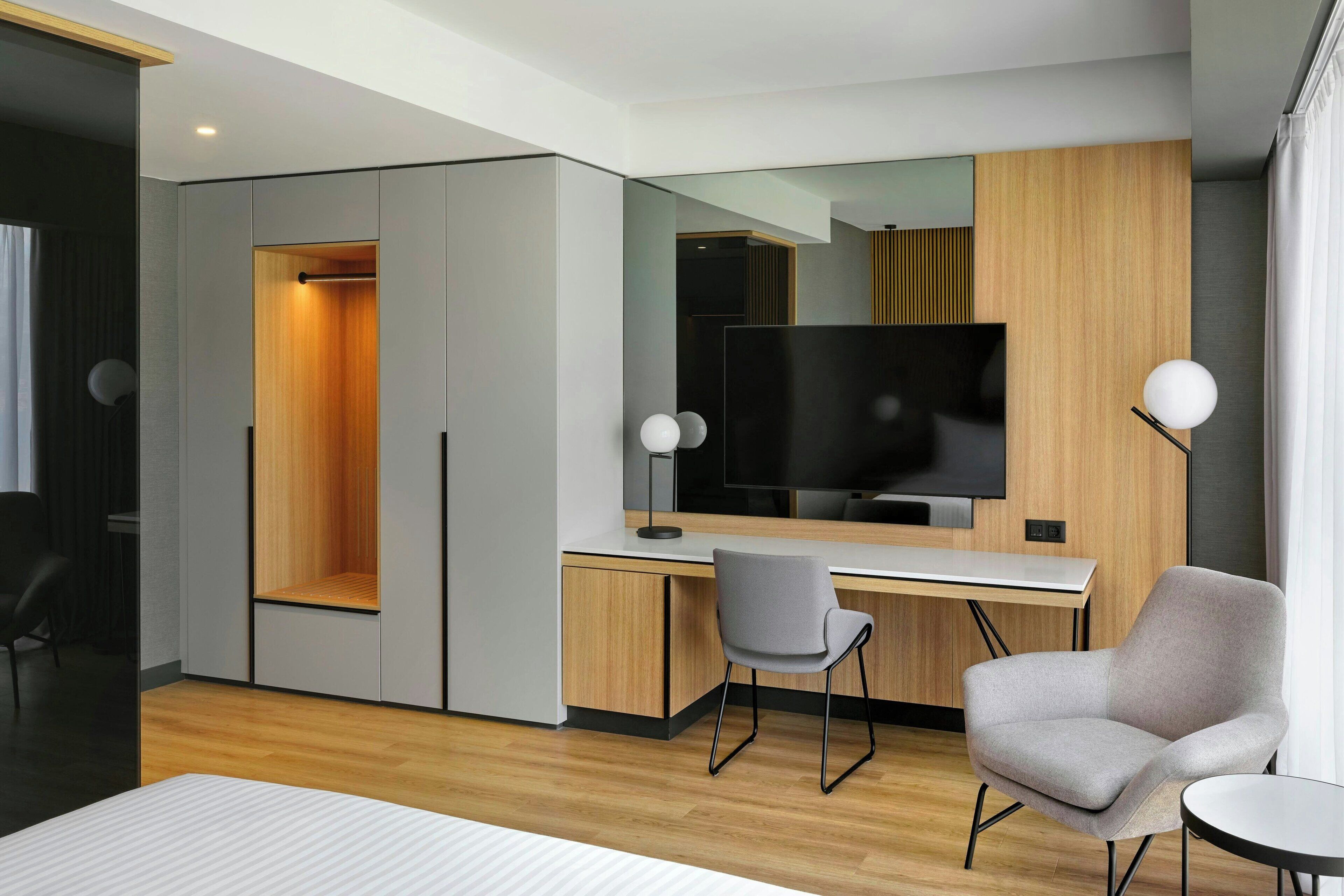 Ein modernes Hotelzimmer mit einem Holztisch, einem grauen Sessel, einem Flachbildfernseher und einem Schrank mit Spiegel.
