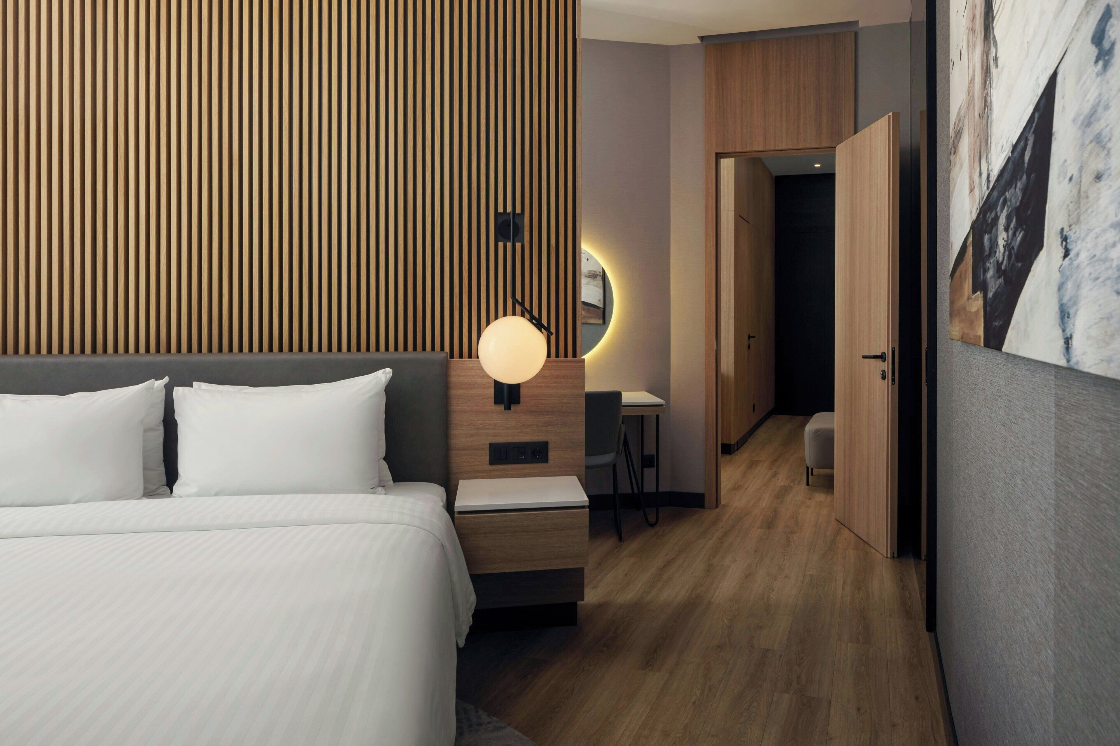 A modern hotel room with a large bed, a desk with a chair, a wall-mounted light, and a door leading to another room.