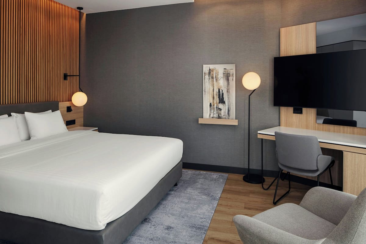 A modern hotel room with a large bed, a desk, a chair, a flat-screen TV, and a wall-mounted artwork.