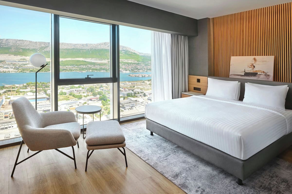 A modern hotel room with a large bed, two armchairs, a small round table, and a view of a lake and mountains.