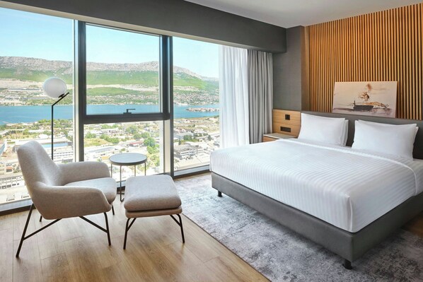 A modern hotel room with a large bed, two armchairs, a small round table, and a view of a lake and mountains.