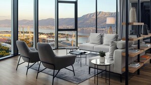 A modern living room with a large window offering a view of a city and mountains. There are two grey armchairs, a sofa, and a coffee table with books and a plant. - AC Hotel by Marriott Split (Split)