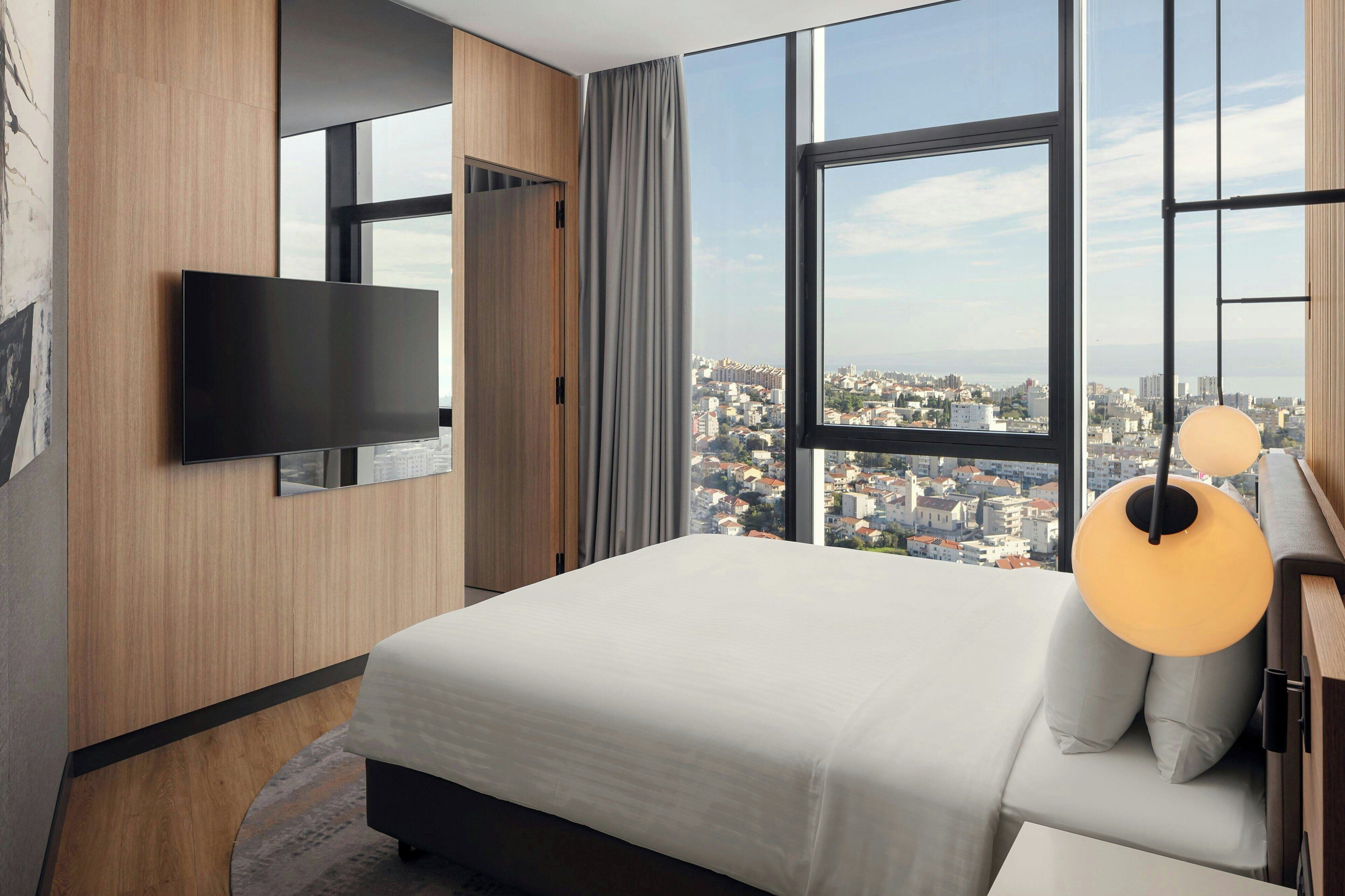 Een moderne hotelkamer met een groot bed, een flatscreen televisie aan de muur, uitzicht over de stad door grote ramen en een moderne lamp.