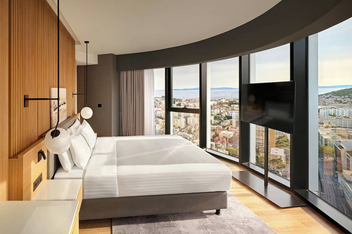 A modern bedroom with a large bed, a TV, and a city view.