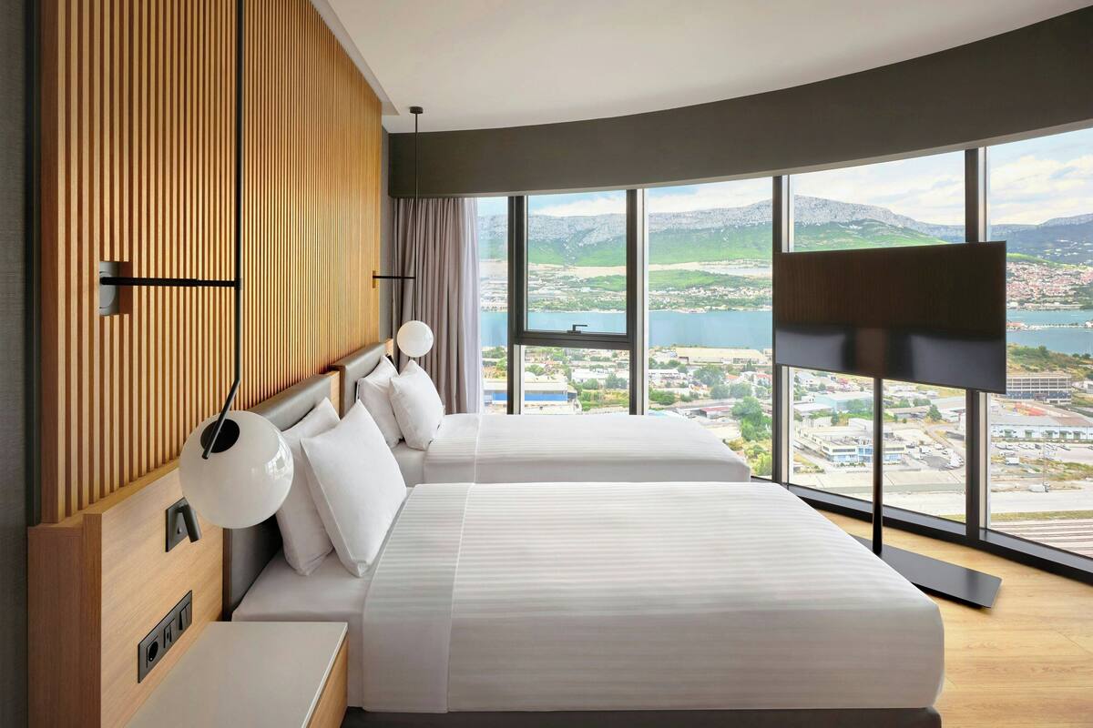 A modern hotel room with two beds, a large window with a city and mountains view, and a wooden panel wall.