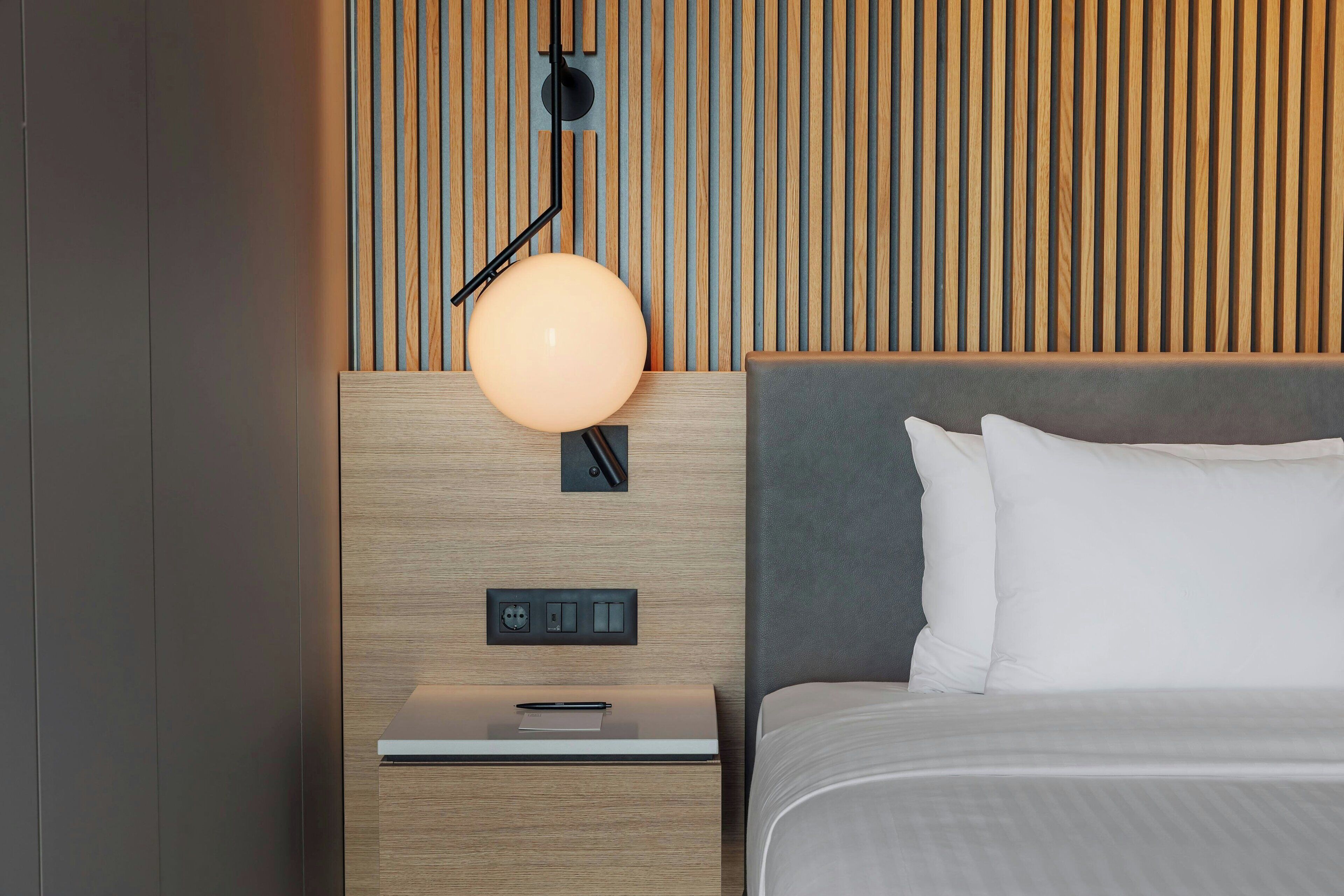 A modern hotel room with a wooden headboard, a bedside table with a lamp, and a bed with white bedding.