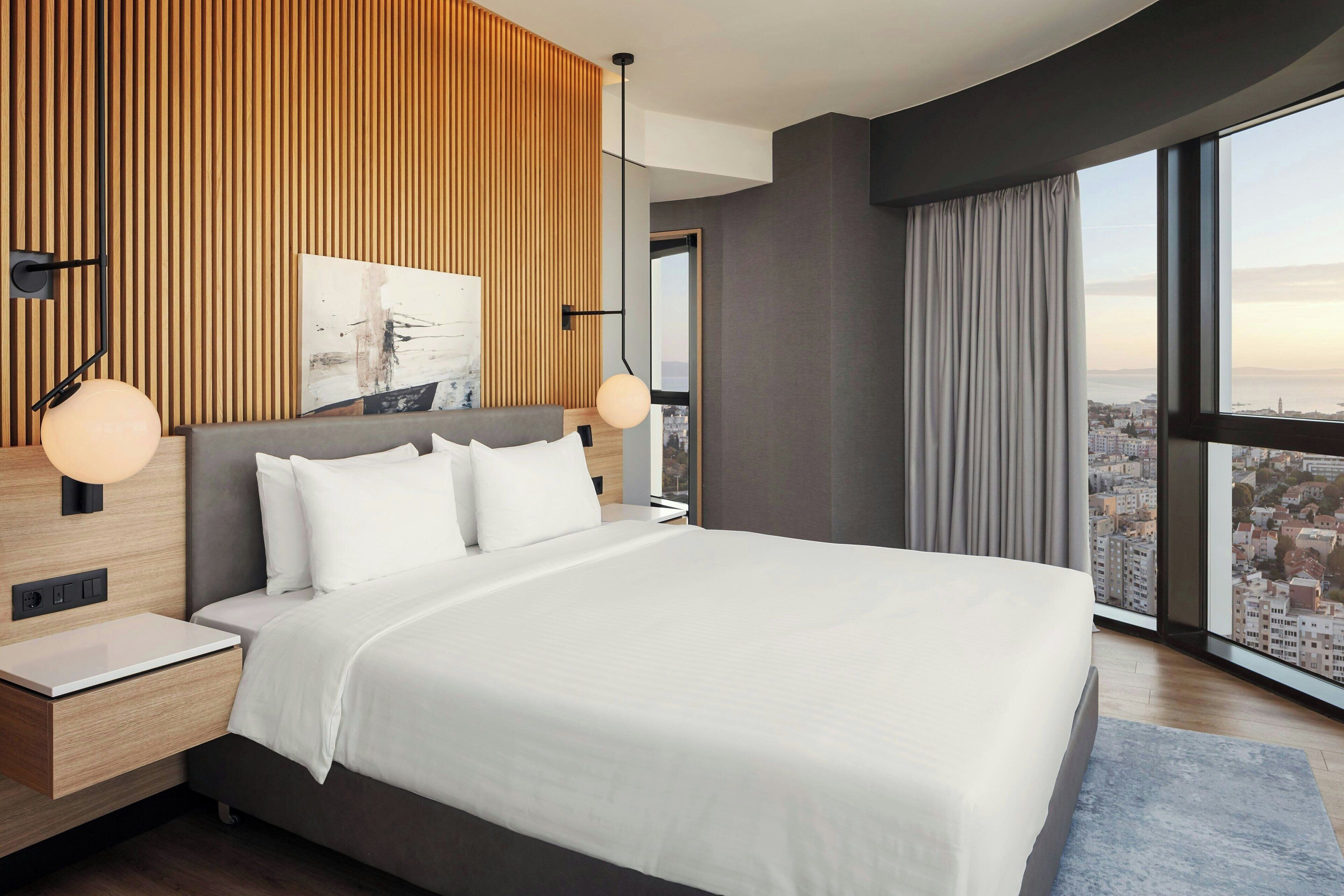 Ein modernes Hotelzimmer mit einem großen Bett, einer Wandpaneinung aus Holz, Blick auf die Stadt und einem Nachttisch mit Lampe.