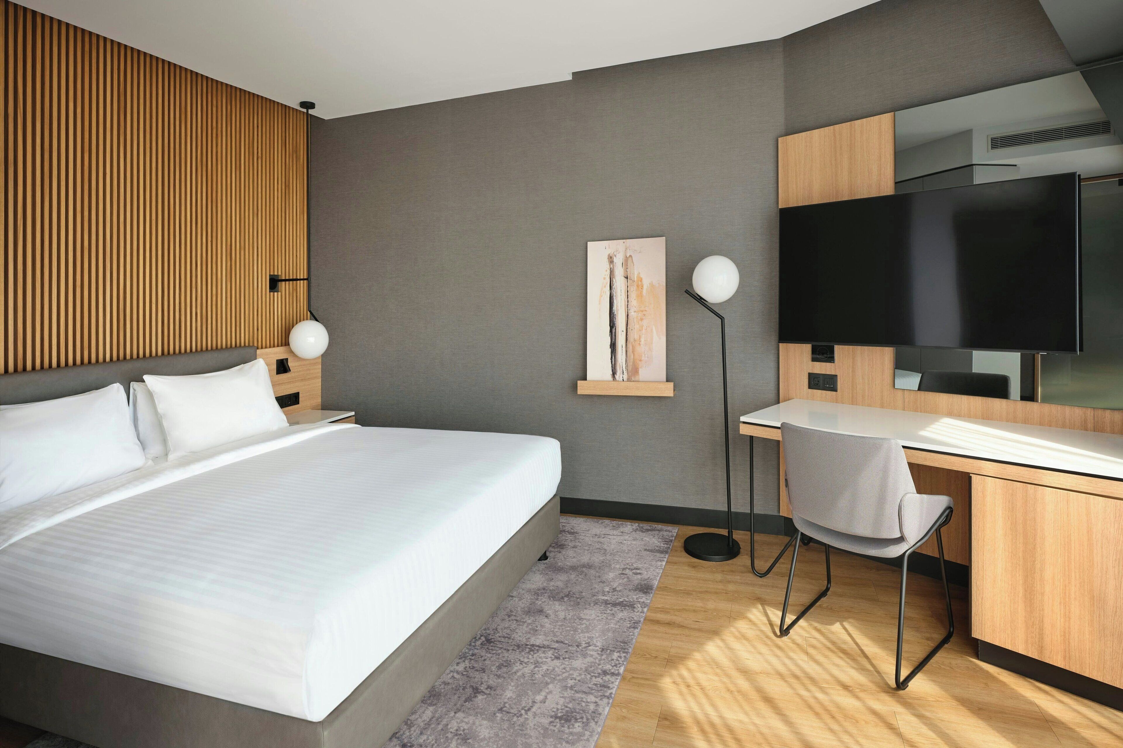A modern hotel room with a large bed, a desk with a chair, a flat-screen TV, and a painting on the wall.