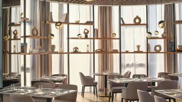 Interior de restaurante moderno com tetos altos, vigas de madeira e janelas amplas. Mesas montadas com pratos, copos e talheres.