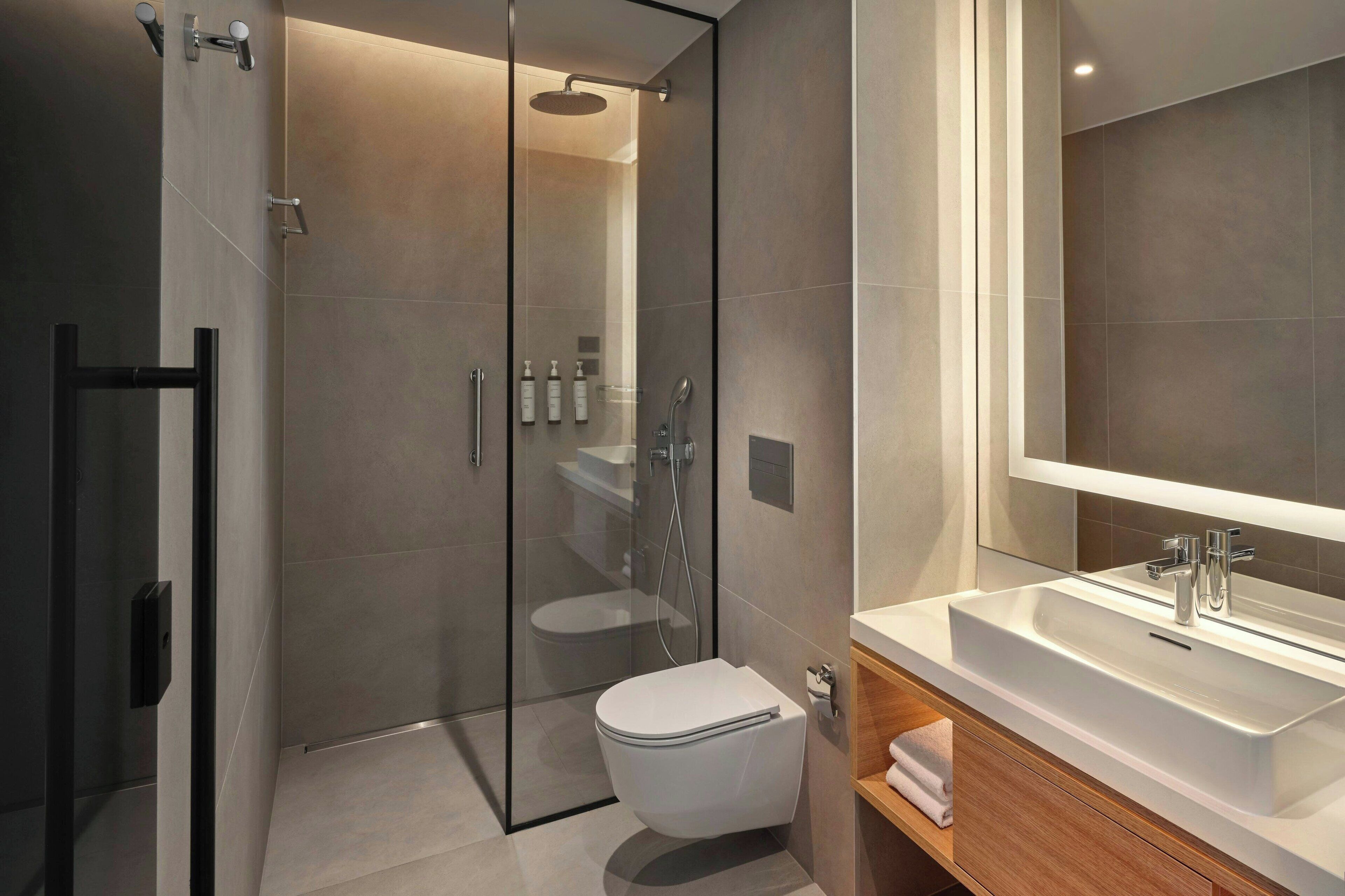 Une salle de bain moderne avec une douche à parois de verre, des toilettes blanches et un grand miroir au-dessus d’un lavabo.