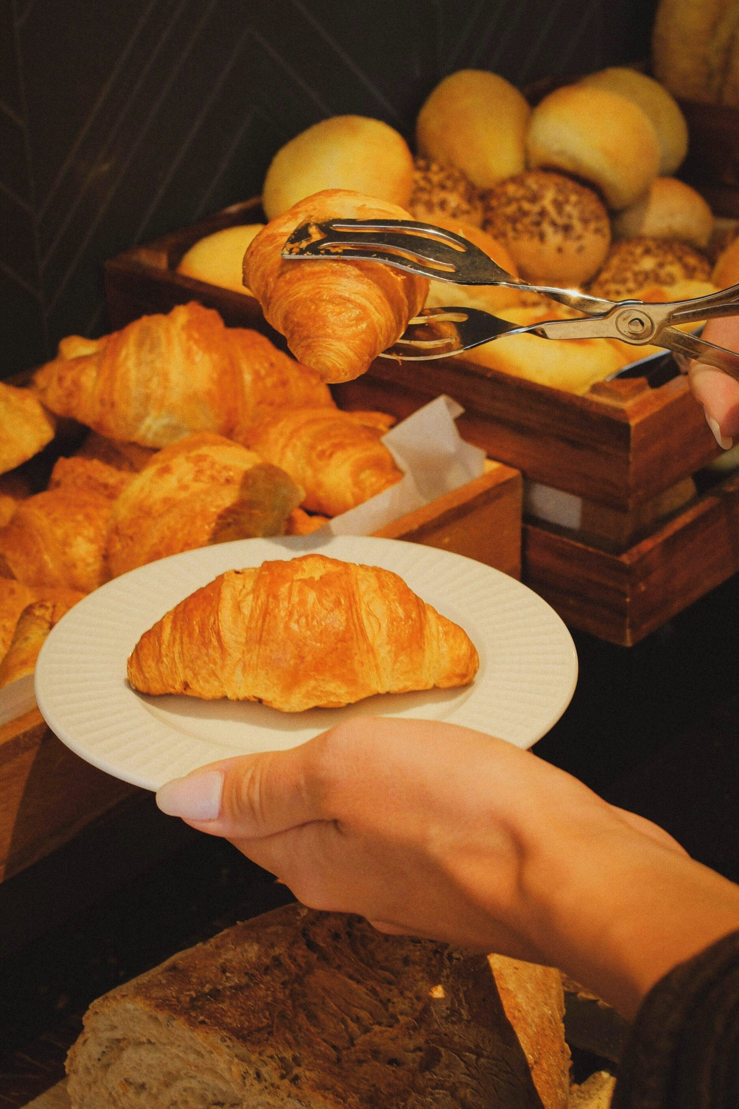 A hand holding a plate with a croissant, with a wooden crate of various baked goods in the background.