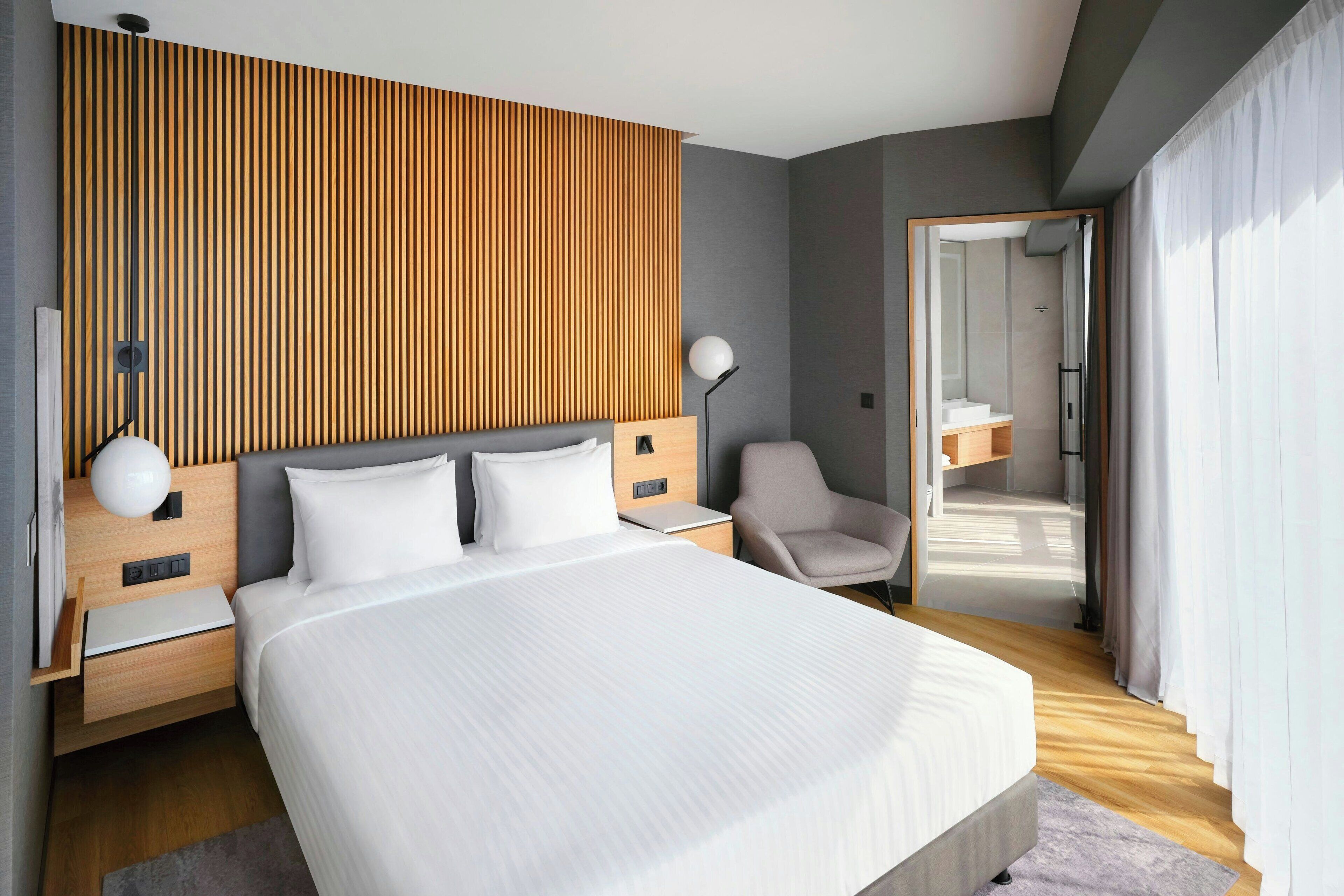 A modern hotel room with a large bed, wooden panel wall, bedside tables, a chair, and a bathroom visible through an open door.