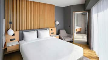 A modern hotel room with a large bed, wooden panel wall, bedside tables, a chair, and a bathroom visible through an open door.