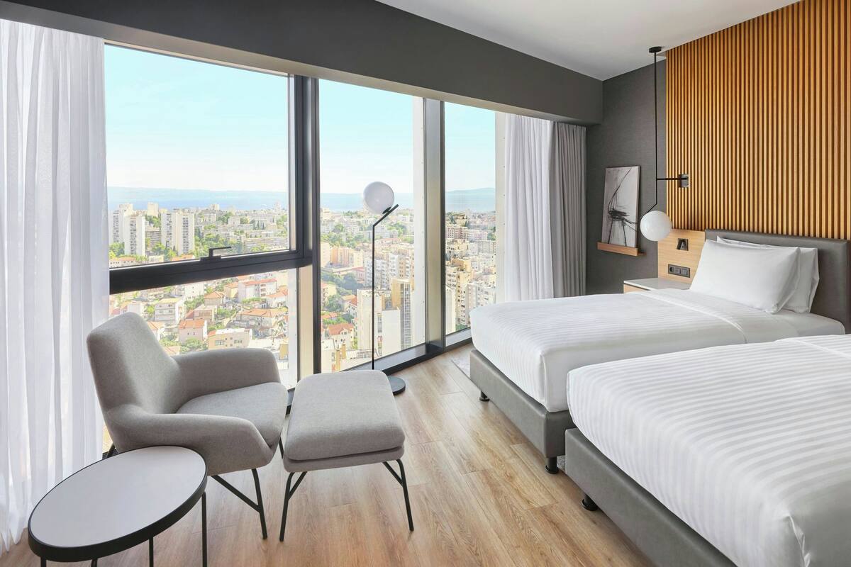A modern hotel room with two beds, a sitting area with a chair and ottoman, a large window with a city view, and a wooden accent wall.