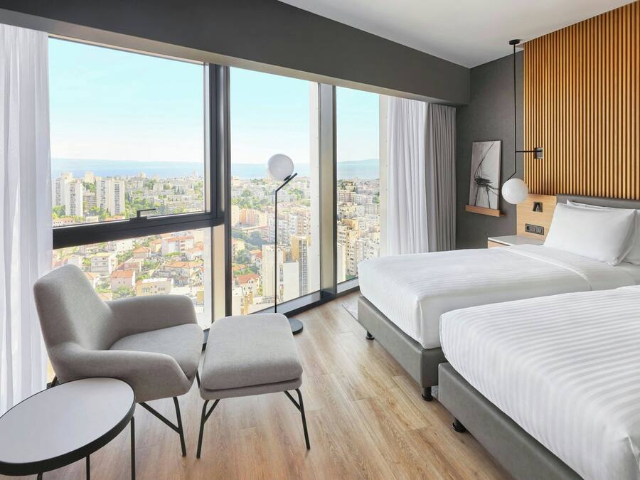 Une chambre d’hôtel moderne avec deux lits, un coin salon comprenant un fauteuil et un pouf, une grande fenêtre donnant sur la ville et un mur décoratif en bois.
