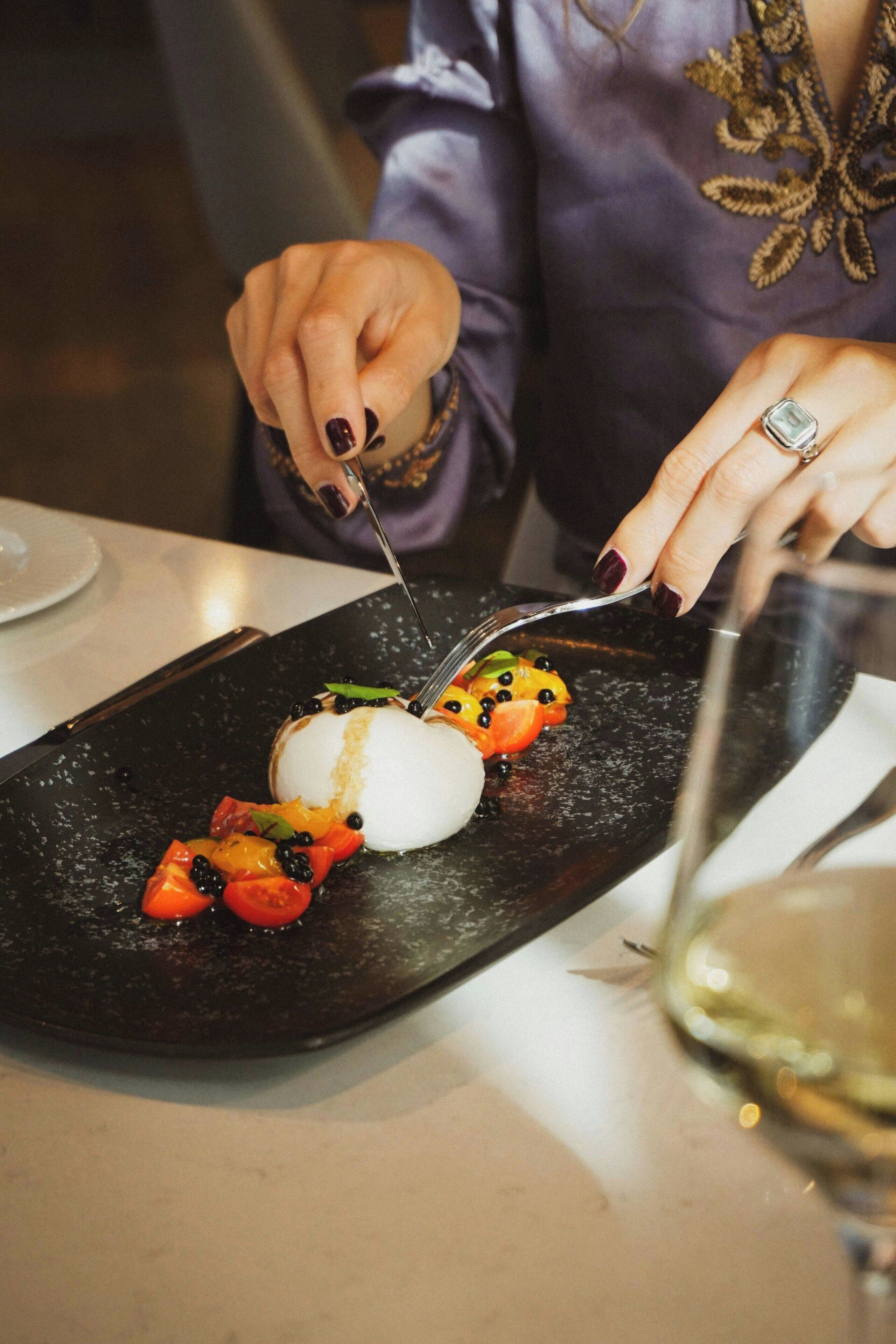 Une personne est en train de découper un dessert sur une assiette noire, avec des fruits et un élément blanc, vraisemblablement un gâteau ou une pâtisserie, dans un restaurant.