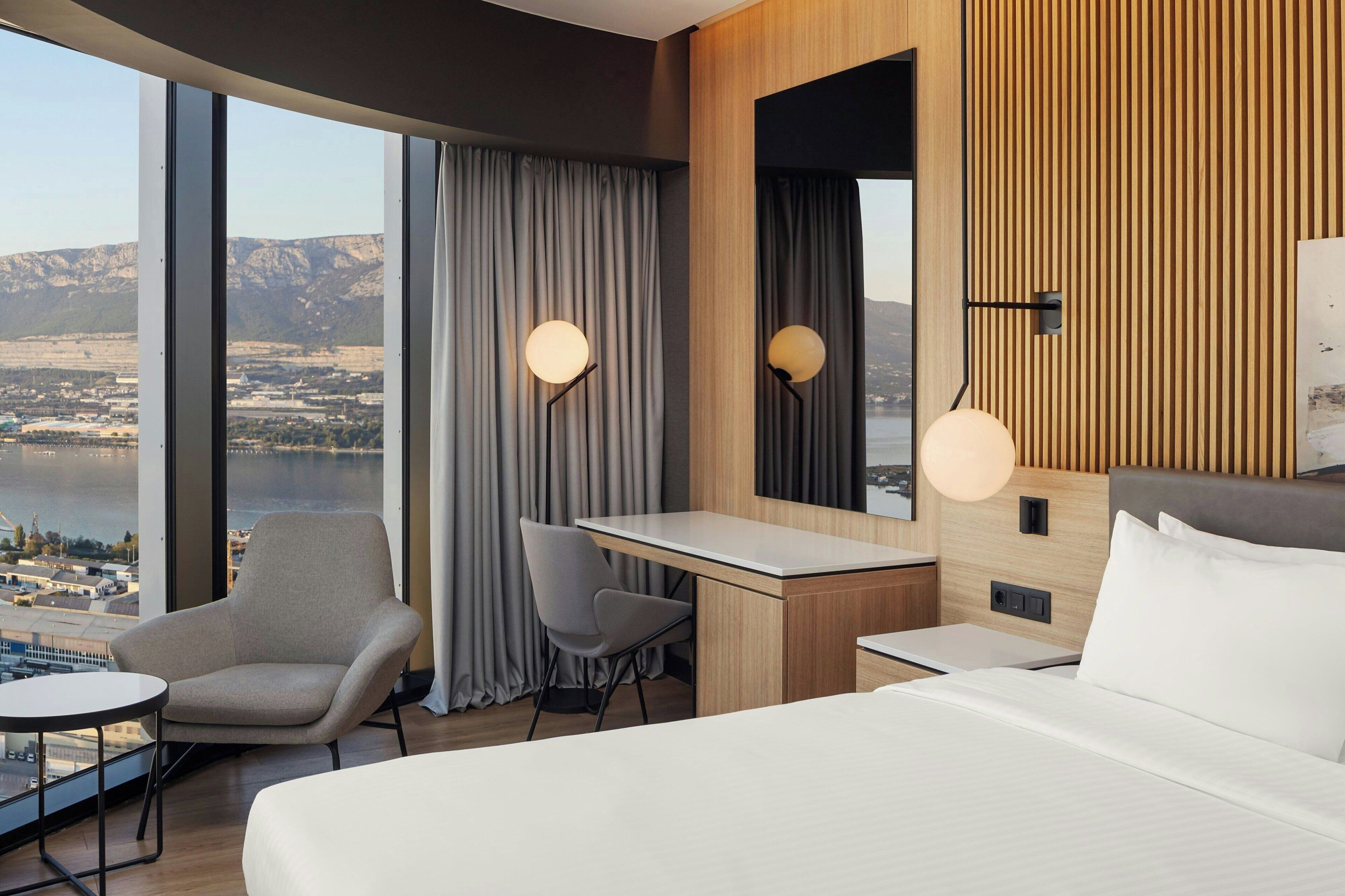 Une chambre d’hôtel moderne dotée d’un grand lit, d’un bureau, d’une chaise et offrant une vue sur un paysage montagneux grâce à de grandes fenêtres.