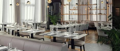 A modern restaurant interior with multiple dining tables, cushioned seating, and pendant lighting.