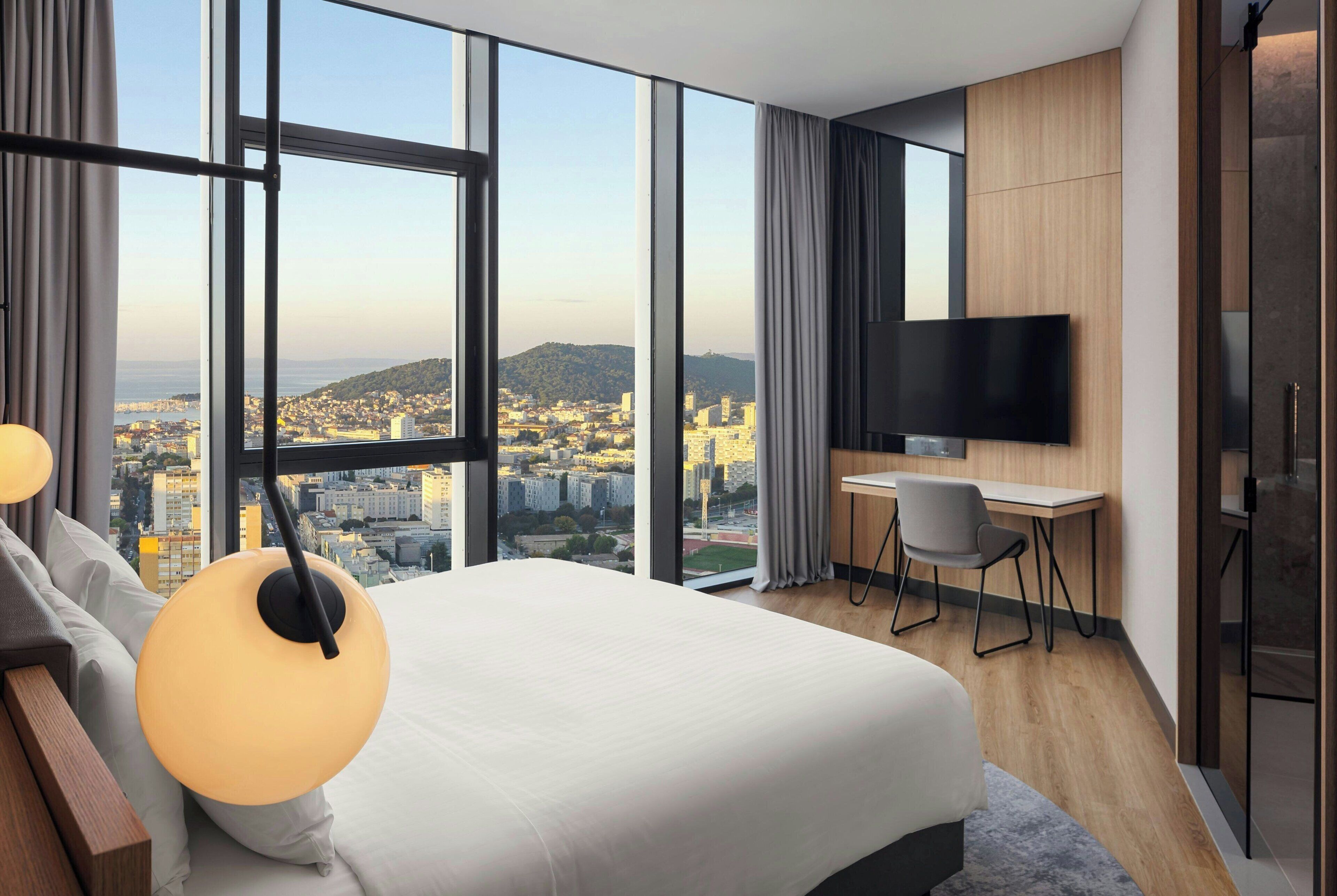 Una habitación de hotel moderna con una cama grande, un escritorio, una silla, un televisor y vistas a un paisaje urbano a través de amplios ventanales.